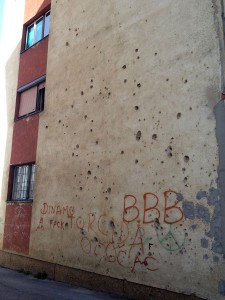 A lateral de um edifícil de quatro andares. Na metade esquerda a parede tem uma flieira vertical de janelas. Na metade direita, uma superfície de concreto cru está repleta de buracos de bala.