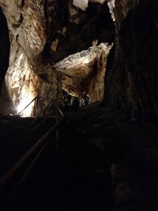 Grande parte da foto é o negro da caverna. Uma figurinha no meio da foto se coloca na frente dos holofotes. A luz apontada para o teto logo acima dá uma ideia da imensidão da caverna.