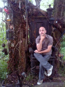 Marcio fazendo cara de inteligente sentado num trono de madeira, na frente de uma árvore, rodeado de lâmpadas e outros objetos inusitados pendurados.
