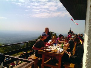 Mesa com seis pessoas almoçando numa varanda. Paisagem de montanhas por trás.